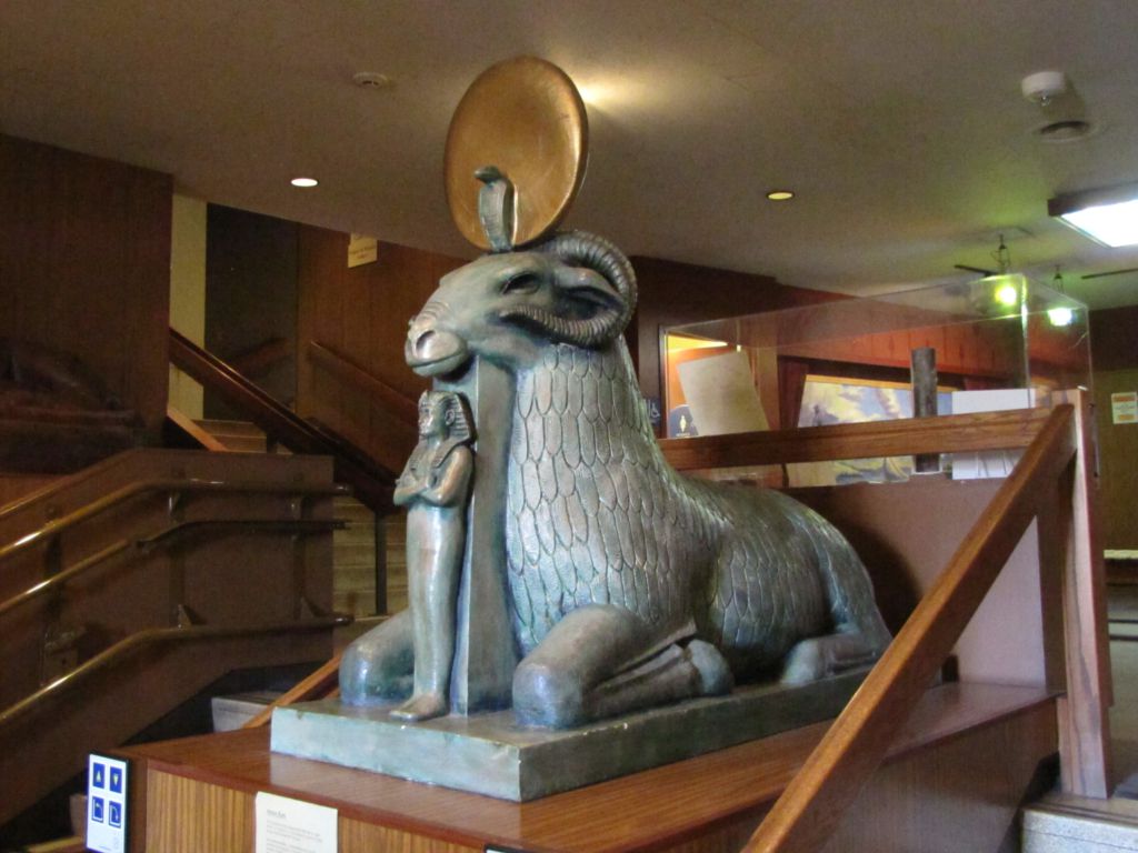 A photo of a Ram statue with human figurine standing just under it's chin